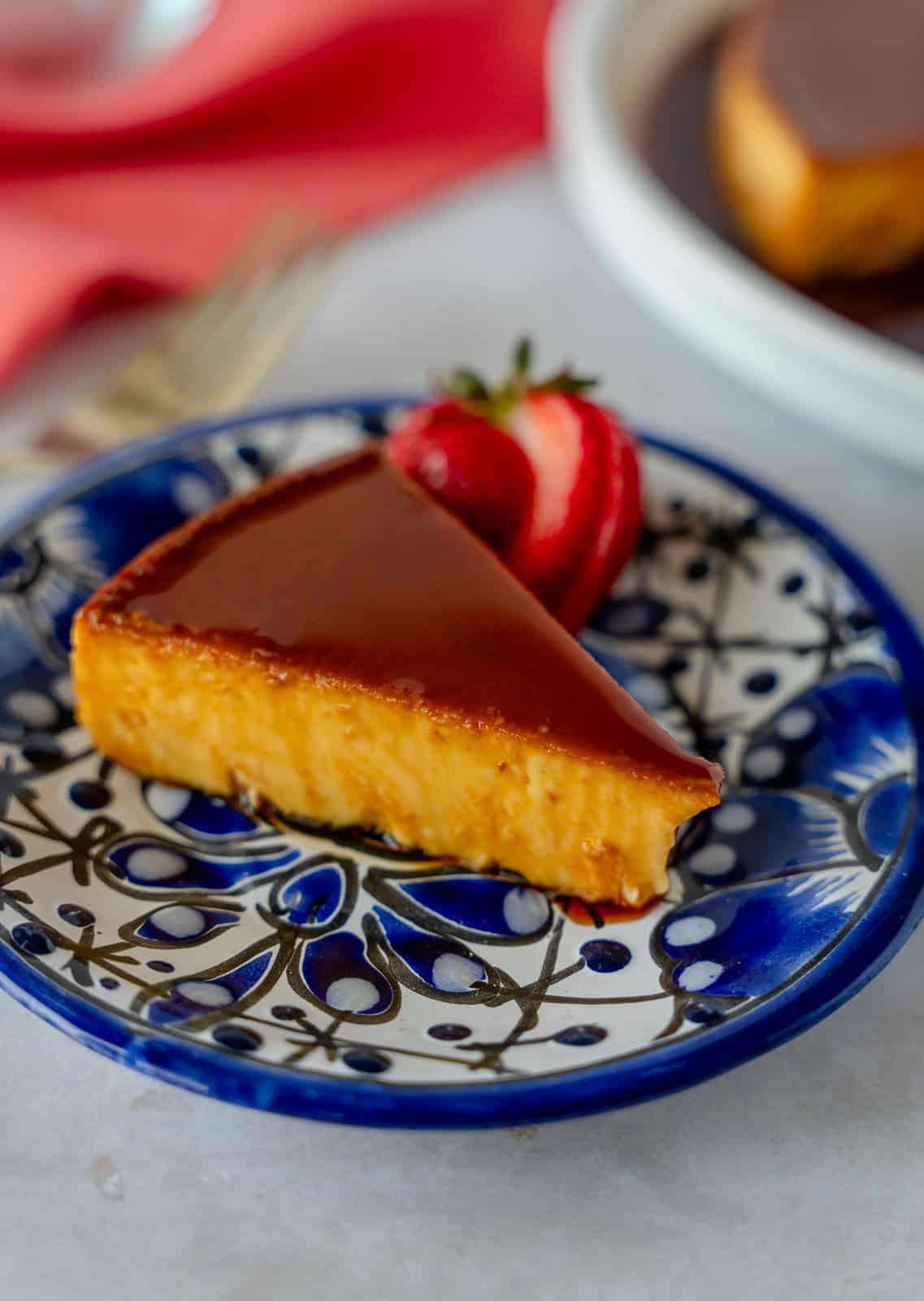 A slice of Mexican flan on a blue plate with more flan in the background.